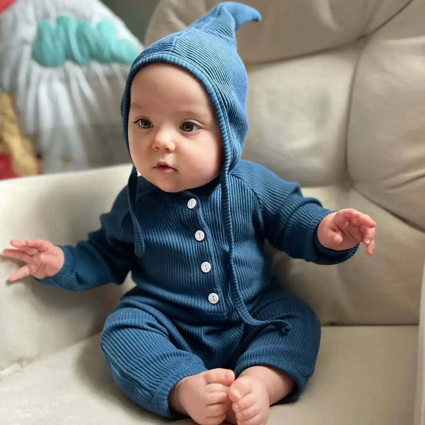 Baby wearing a blue knitted outfit and hat sitting on a beige couch.