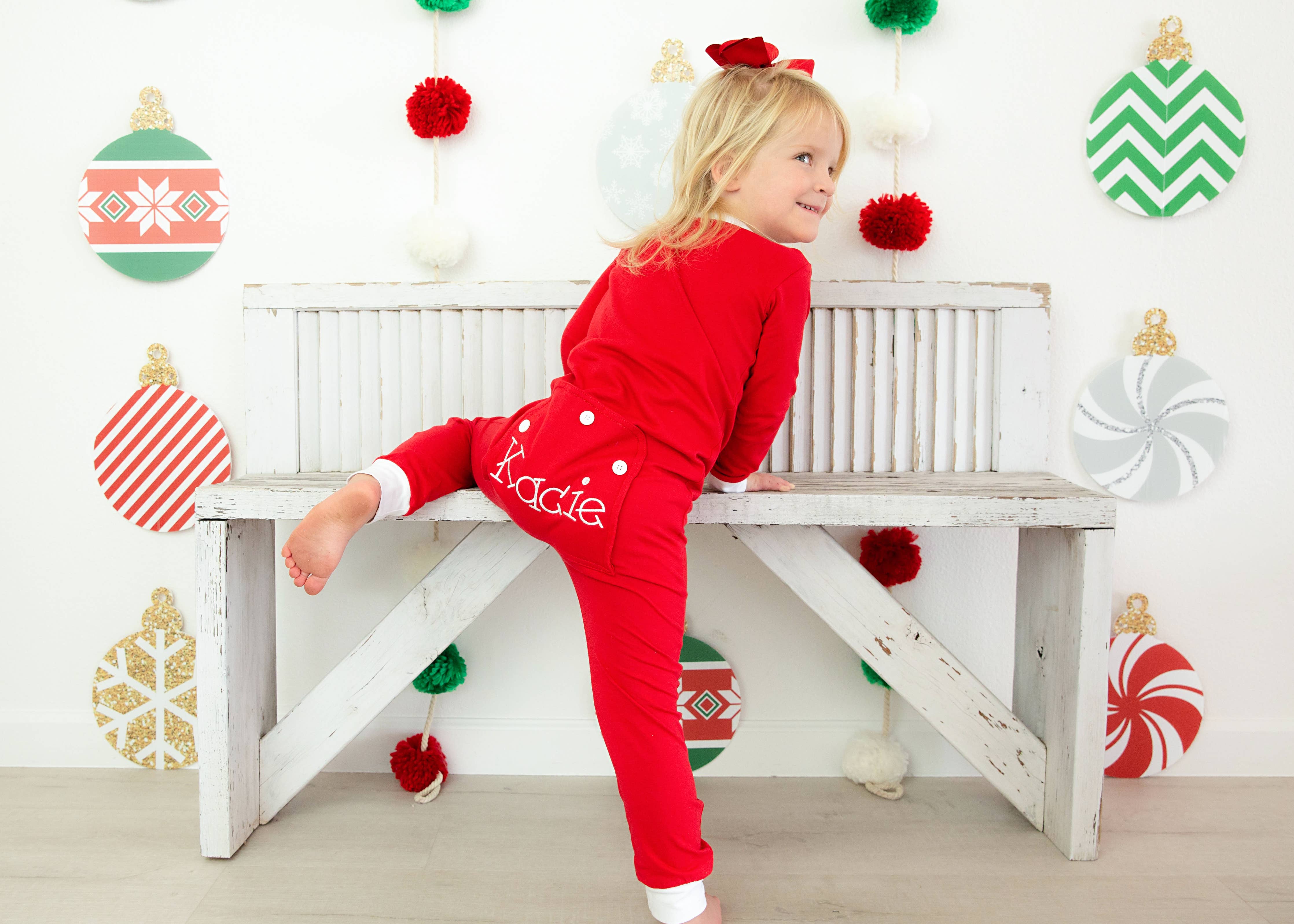 Child in red pajamas with 'Katie' on a white bench decorated with Christmas ornaments.