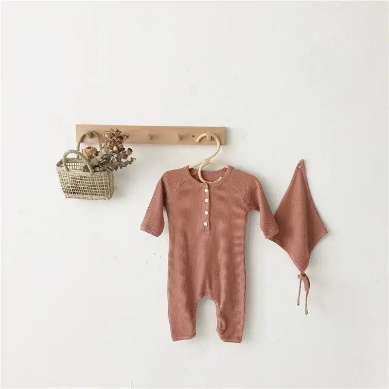 Brown baby romper and hat hanging on a wooden hanger against a white wall.