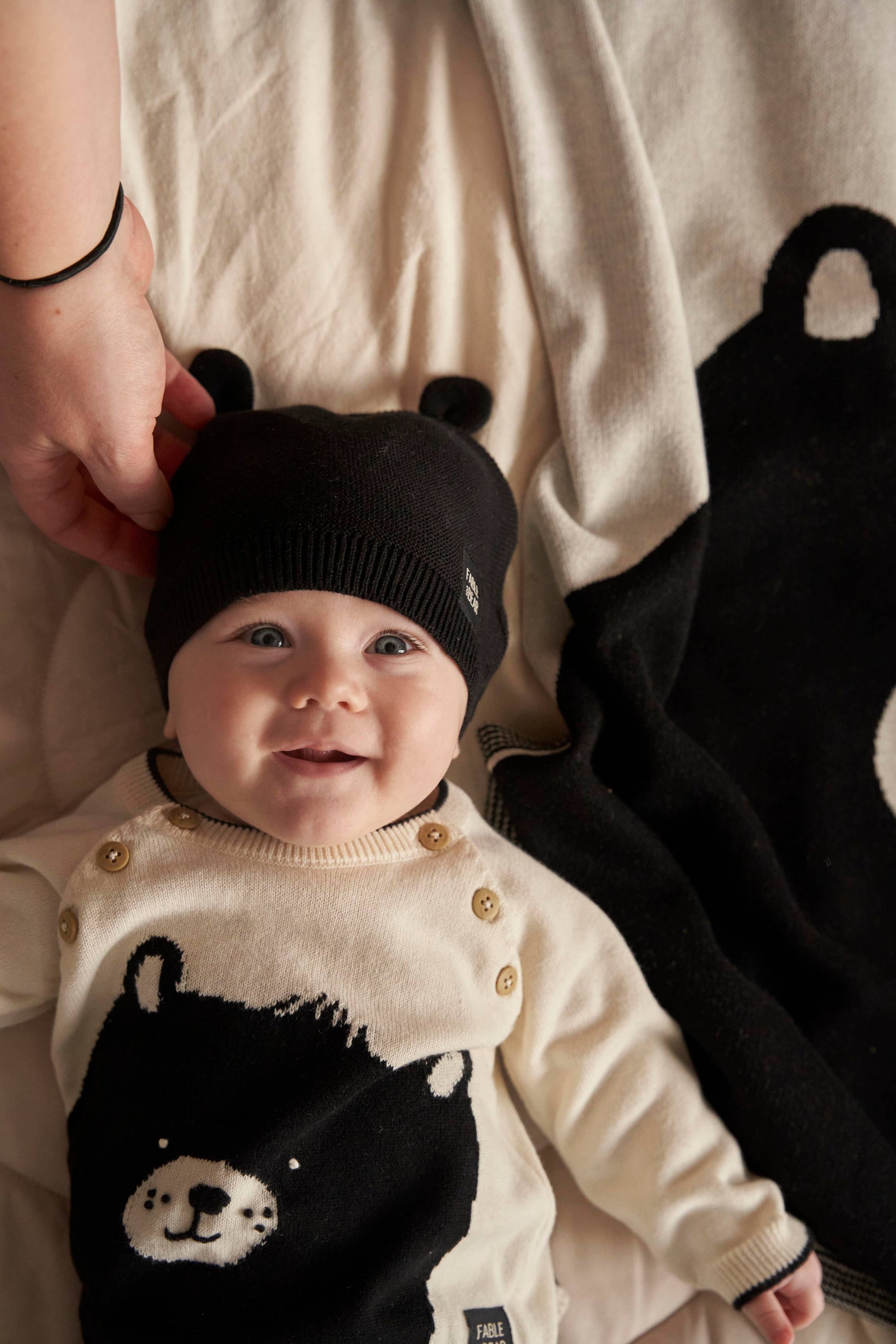 Baby wearing a black and white outfit with bear design and a matching hat, standing next to a person.