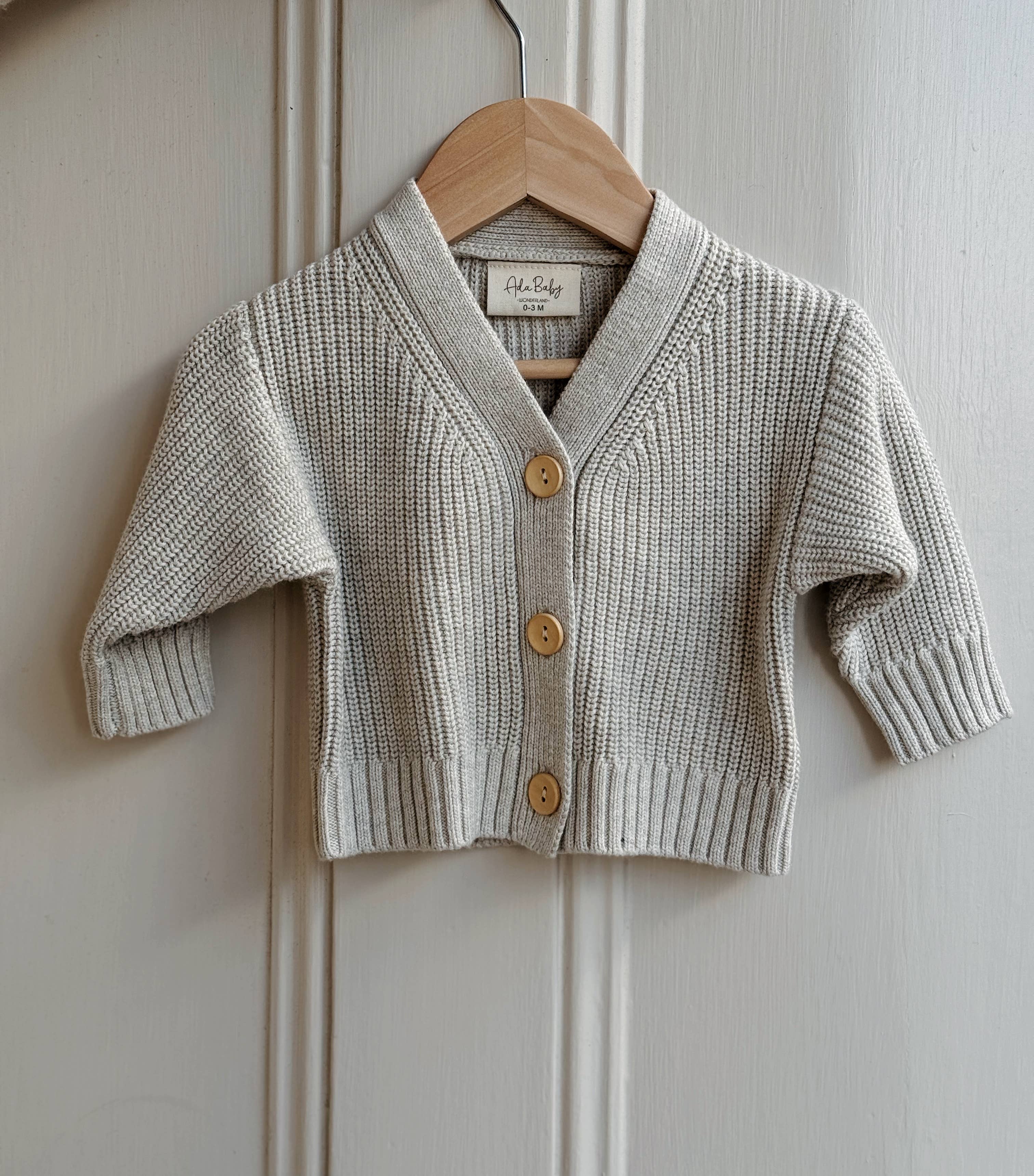 Baby cardigan with buttons on a wooden hanger against a white paneled wall