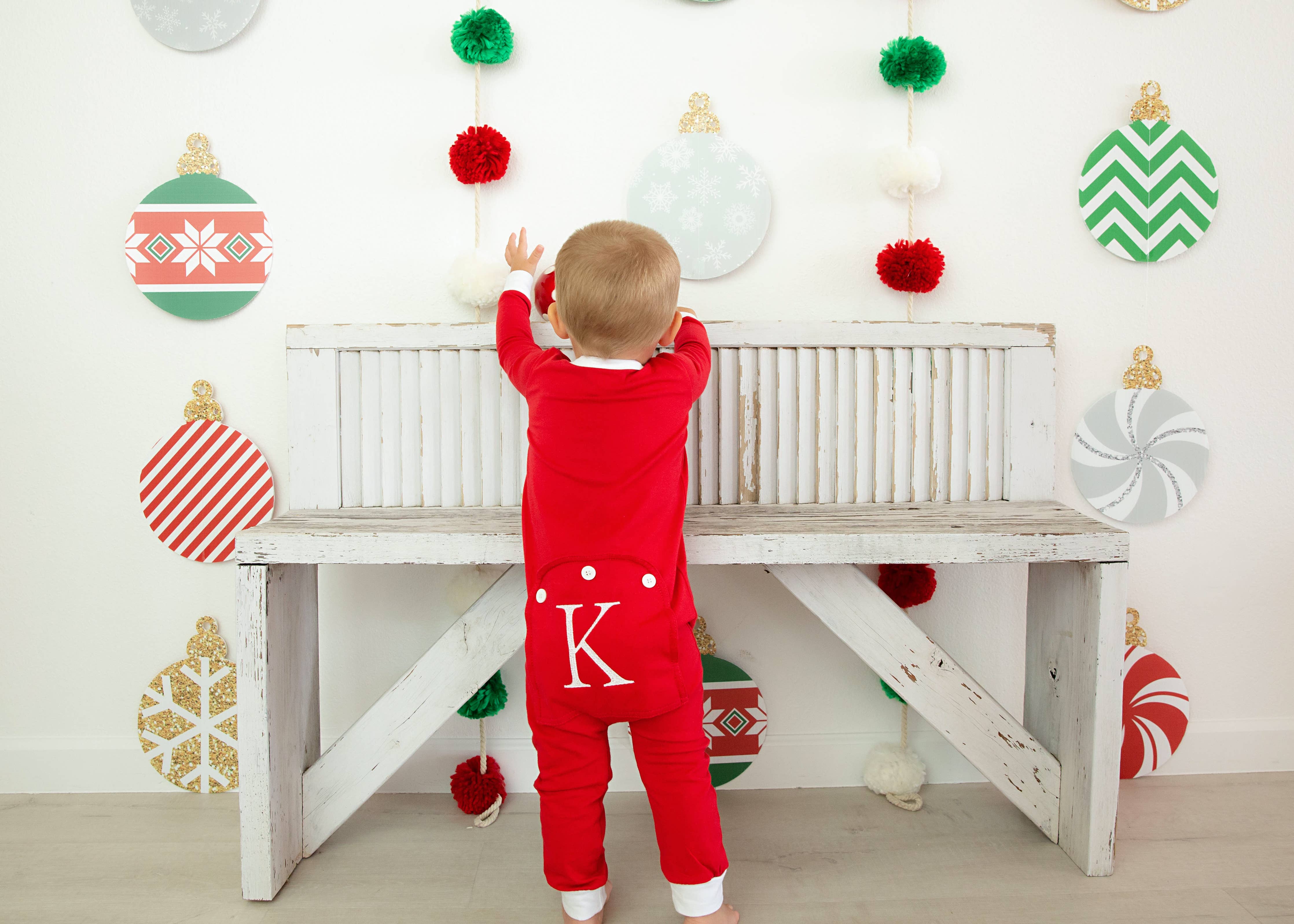 Child in a red onesie with a white letter 'K' standing in front of a wall decorated with festive ornaments.