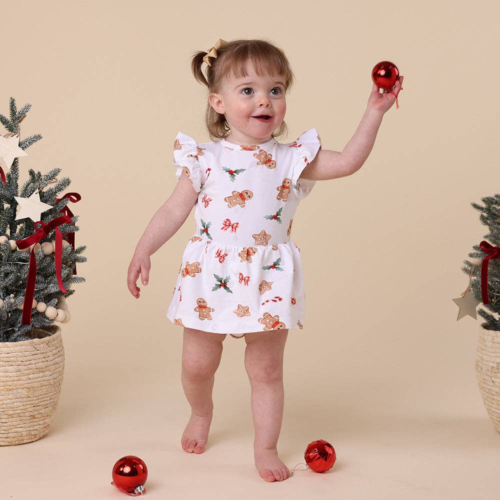 Child in a white dress with gingerbread men pattern holding a red ornament against a beige background.