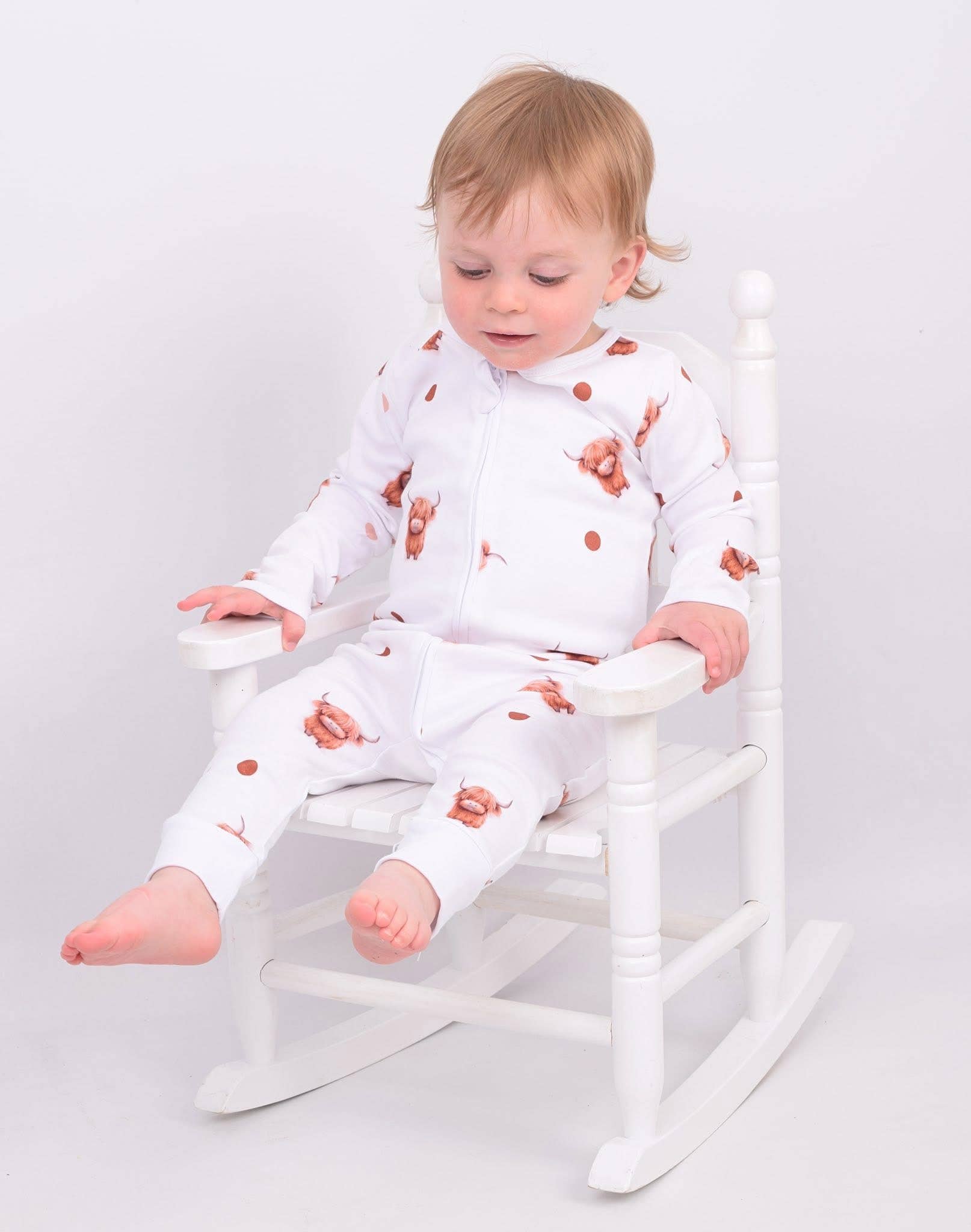 Child wearing a white outfit with highland cow patterns sitting on a white chair against a plain background