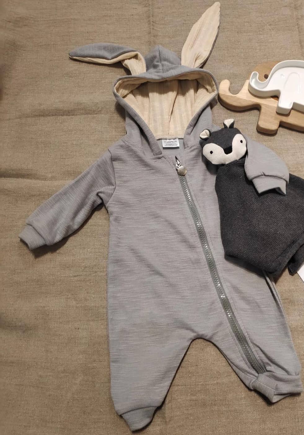 Gray baby sleepsuit with bunny ears and tail, next to a plush toy on a brown surface