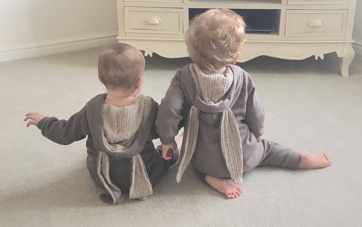 Two children in matching outfits with scarves sitting on a light-colored floor.