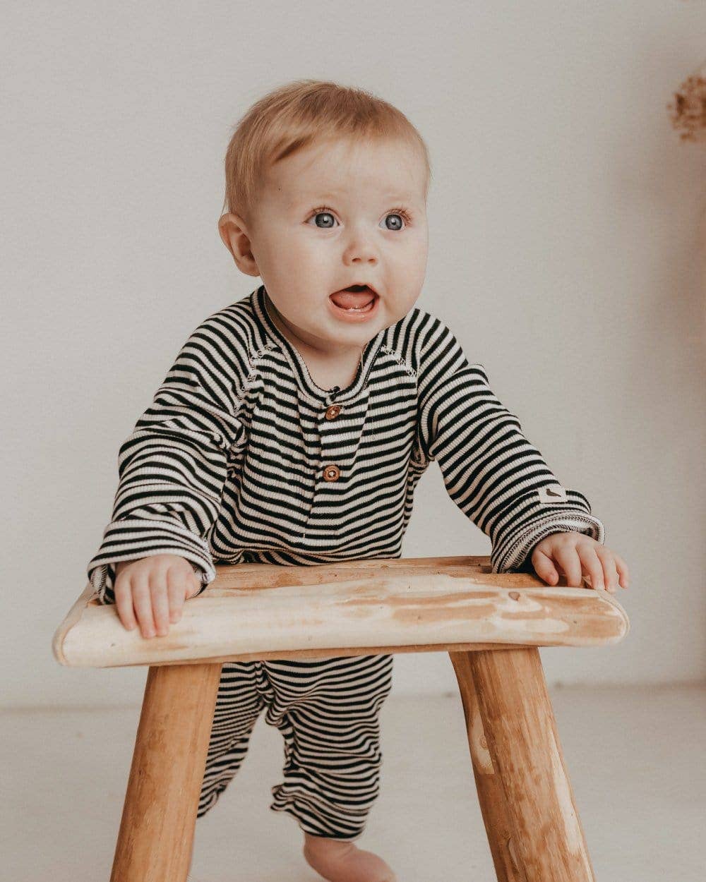 Baby in a striped rib playsuit sitting on a wooden stool against a plain background