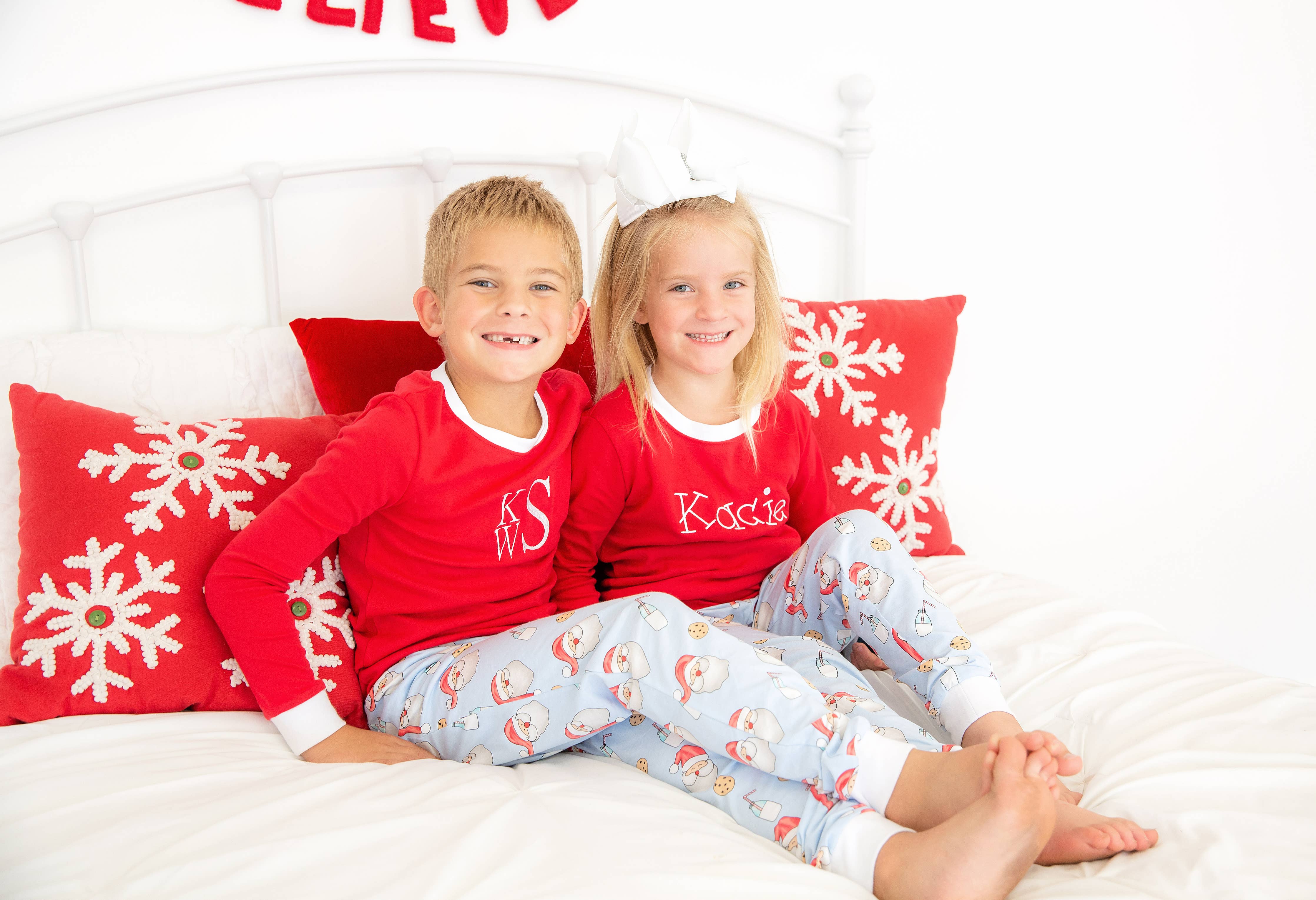 Two children in red pajamas with monograms sitting on a bed with snowflake pillows.
