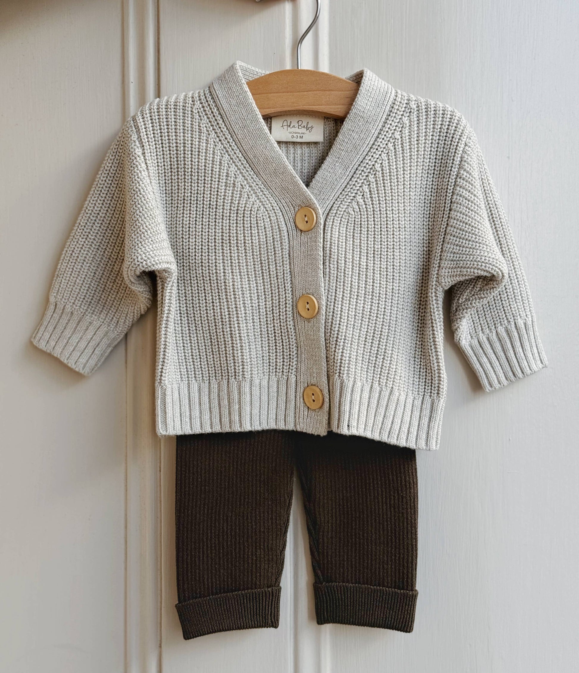 Children's melange knit cardigan with button details and brown pants on a hanger against a white background.