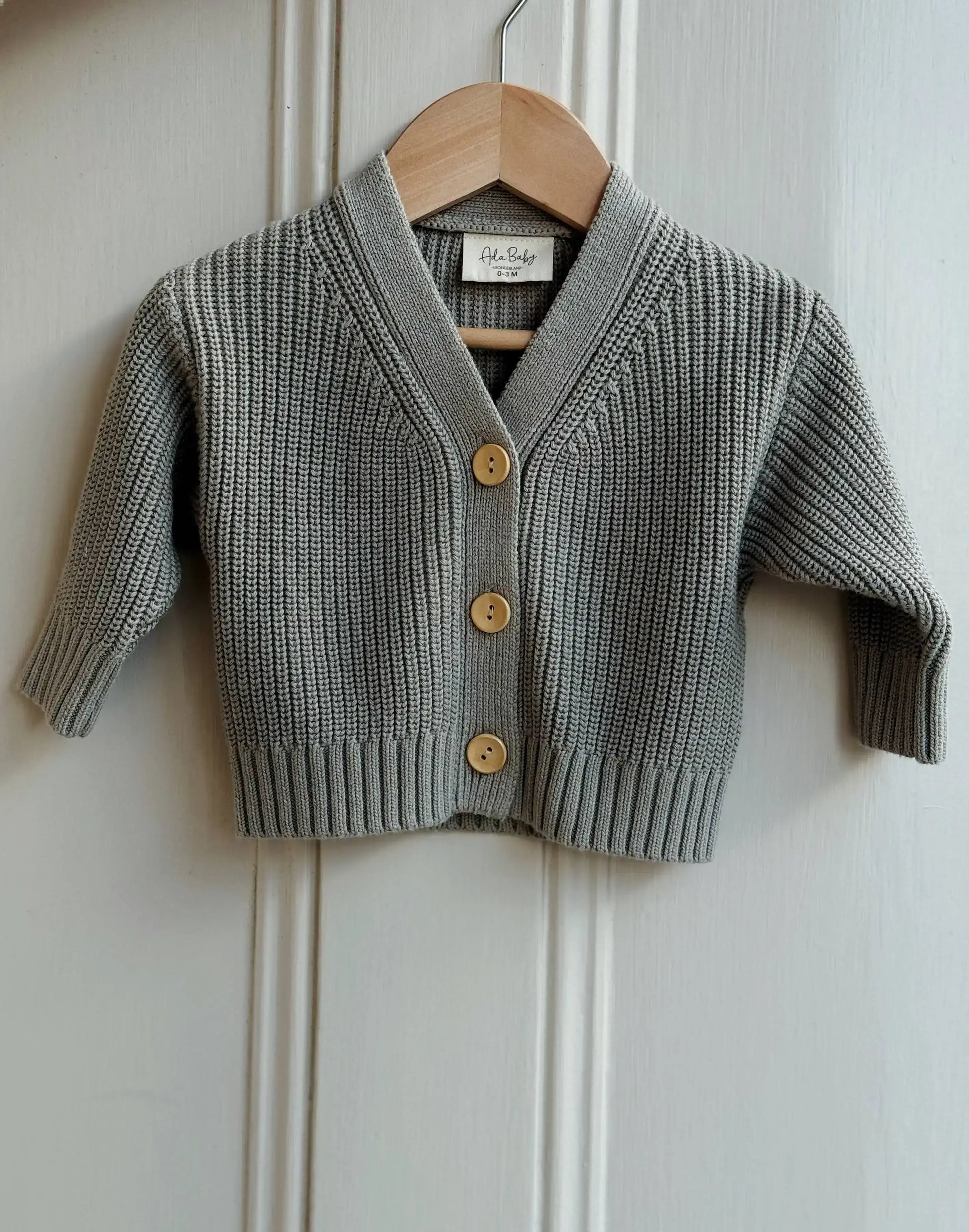 Light green knitted cardigan with yellow buttons on a wooden hanger against a white paneled wall.