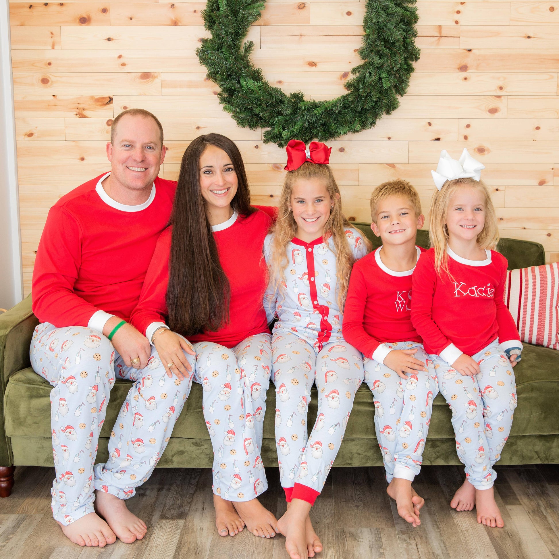 Family of five wearing matching red and white pajamas sitting on a couch with a Christmas wreath in the background.