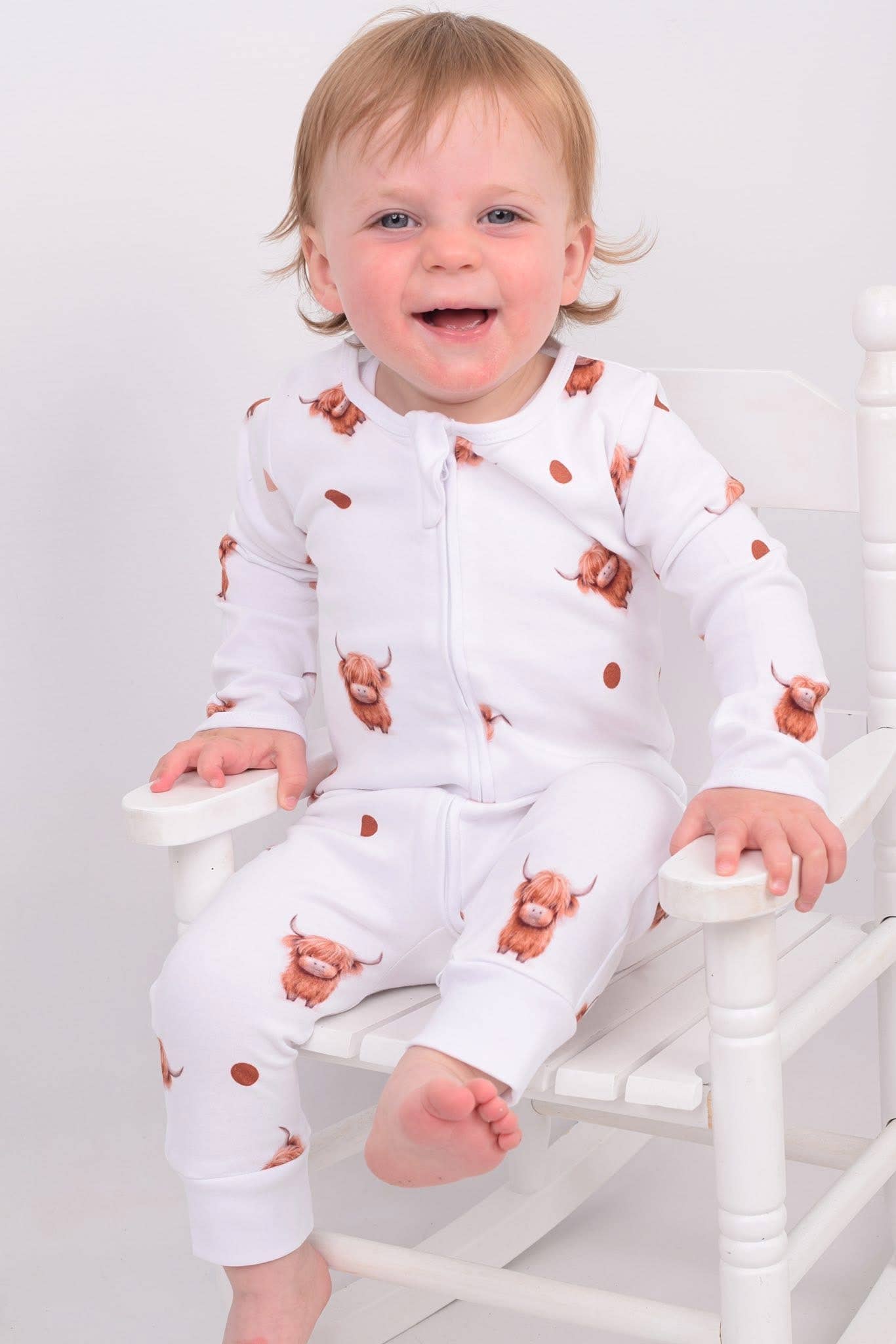Baby wearing a white sleepsuit with brown animal prints, sitting on a white chair.