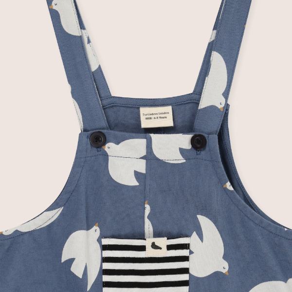 Blue dungaree with white bird pattern and black and white striped pocket on a light gray background