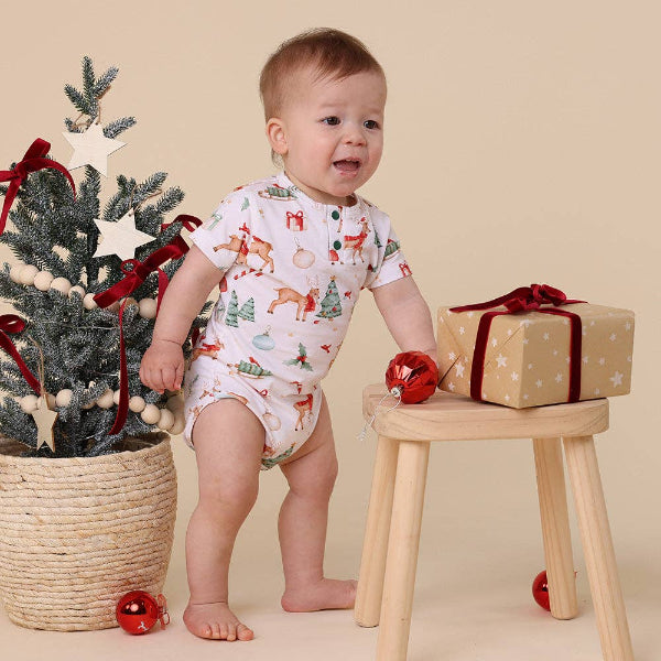 Baby in a festive romper standing next to a Christmas tree and gift box on a beige background