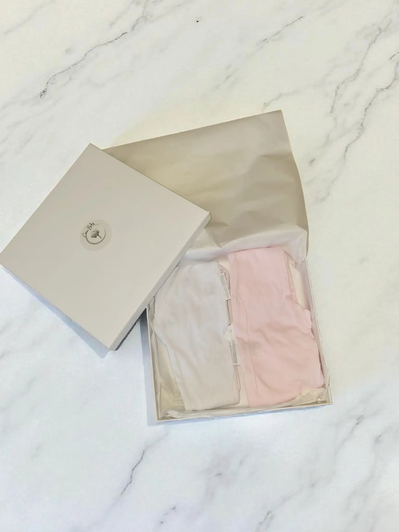 Folded pairs of baby cotton leggings in white and pink, presented in an open Cotton Baby gift box on a marble background