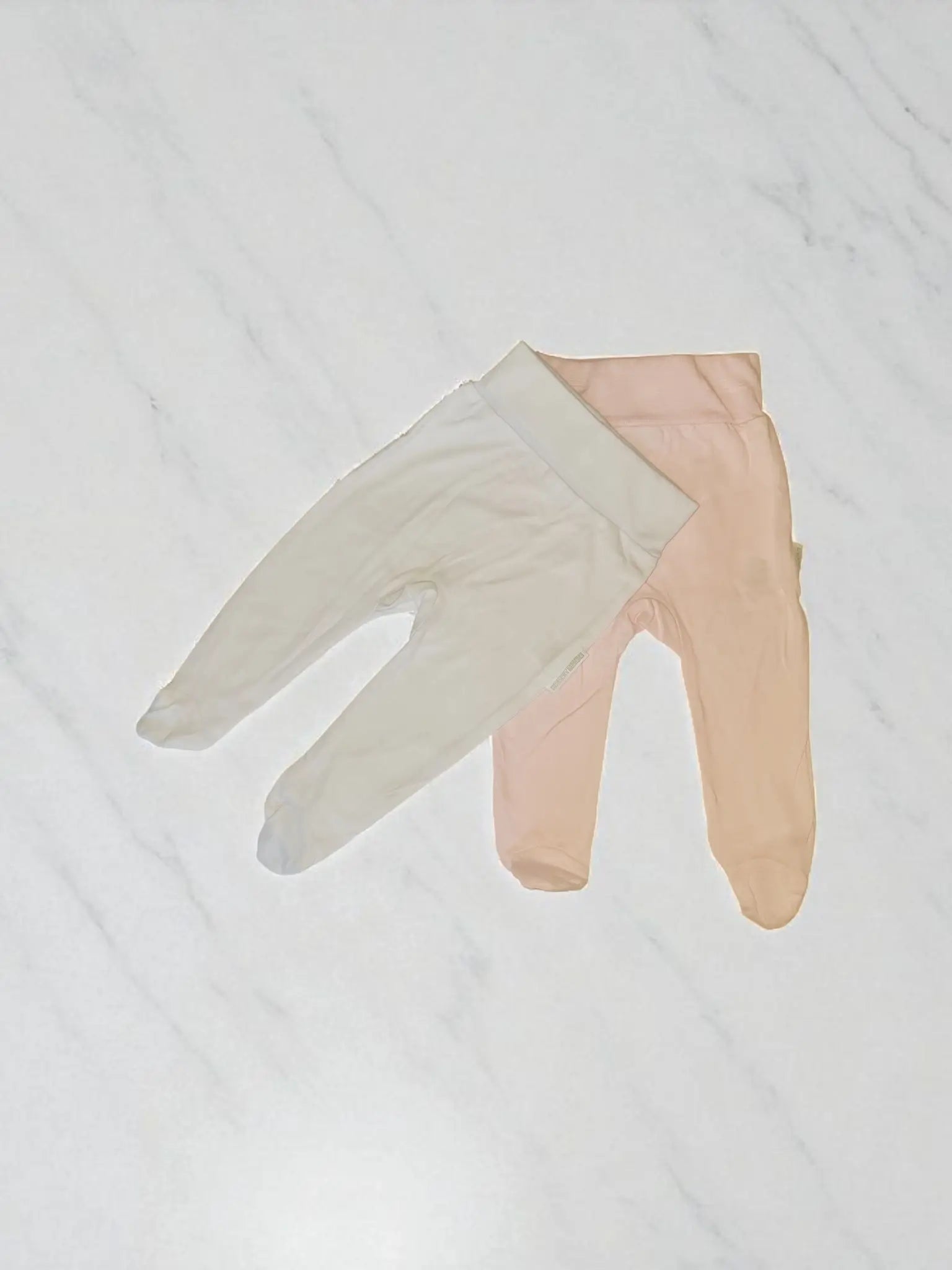 Two pairs of cotton baby leggings with feet, one in white and one in pink, laid flat on a white marble background