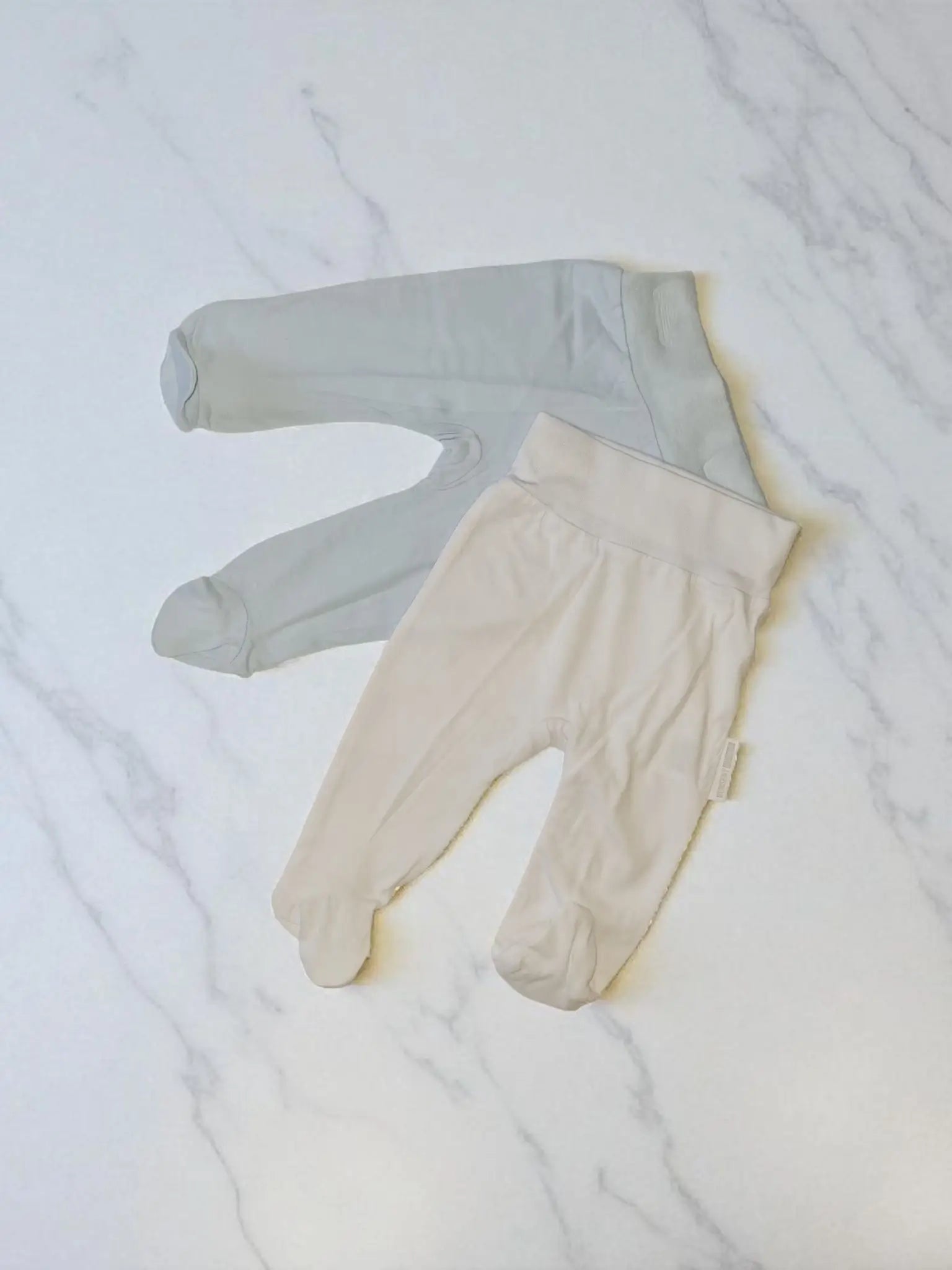 Two pairs of soft cotton baby leggings with feet, one in sky blue and one in white, displayed on a white marble surface