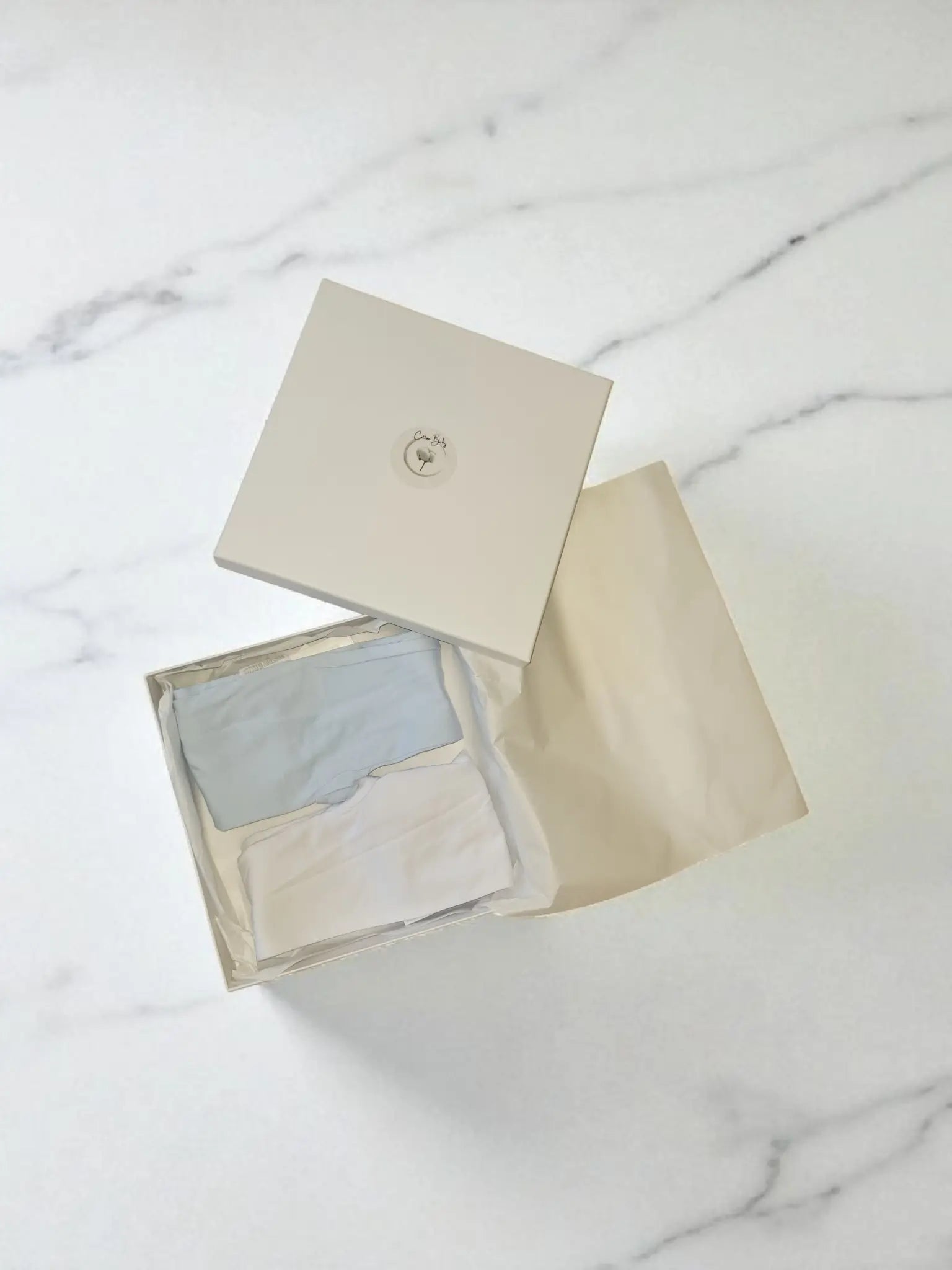 Two pairs of soft cotton baby leggings with feet, one in sky blue and one in white, neatly folded inside a Cotton Baby gift box on a white marble surface