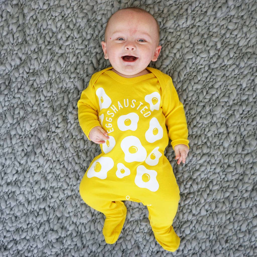 Baby wearing a yellow sleepsuit with white text and patterns, lying on a textured gray surface.