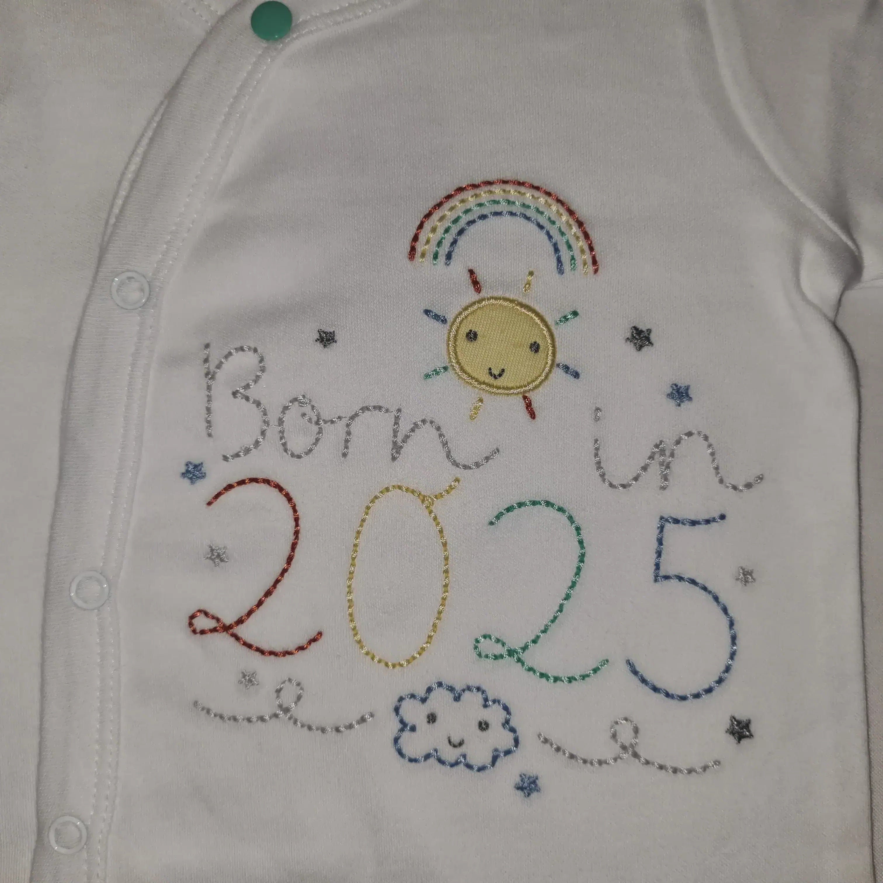 Close-up front detail of a white cotton baby grow with colourful embroidery featuring ‘Born in 2025’, rainbow, sun, stars, and cloud design, with snap button fastening, on a white background