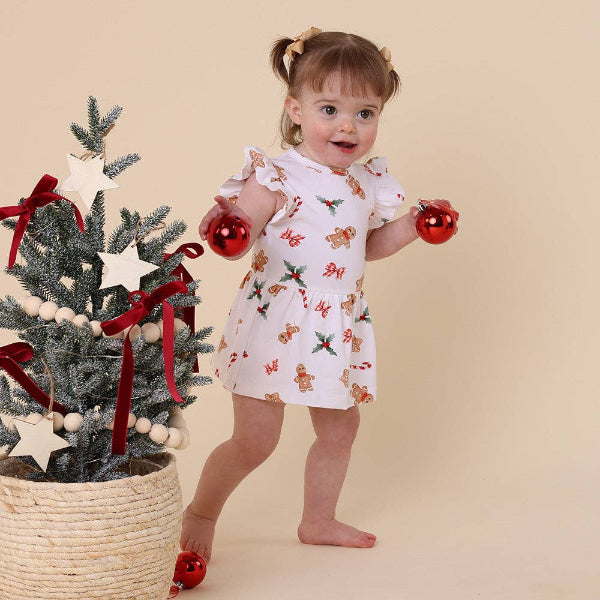 Child in a festive dress holding Christmas ornaments next to a small decorated tree on a beige background