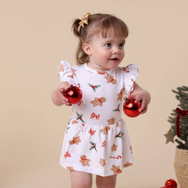 Child wearing a festive dress with gingerbread men and holly patterns, holding Christmas ornaments against a beige background.