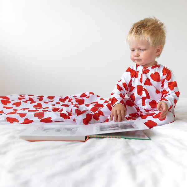 Child wearing a red and white heart-patterned onesie sitting on a bed with a book.