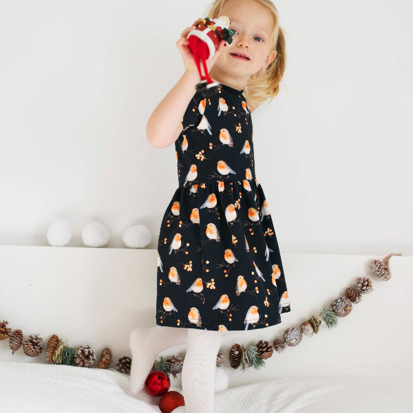 Child wearing a black dress with bird pattern, sitting on a white surface with Christmas decorations.