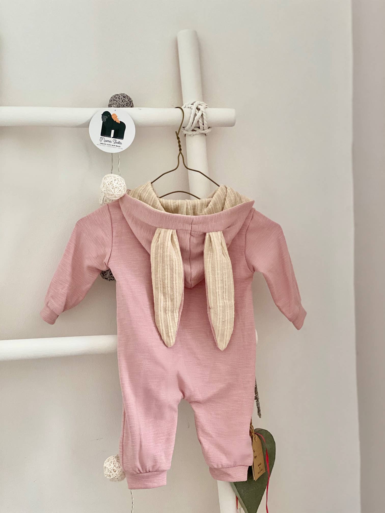 Pink baby sleepsuit with bunny ears on a hanger against a white wall.