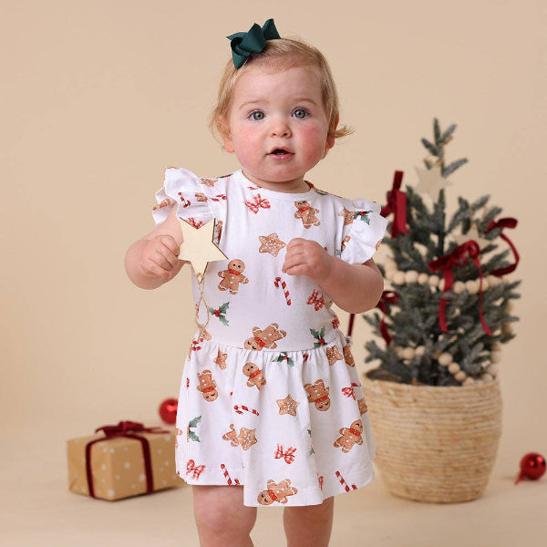 Baby in a Christmas-themed dress with gingerbread men and candy cane pattern, standing in front of a small decorated tree and gift box.