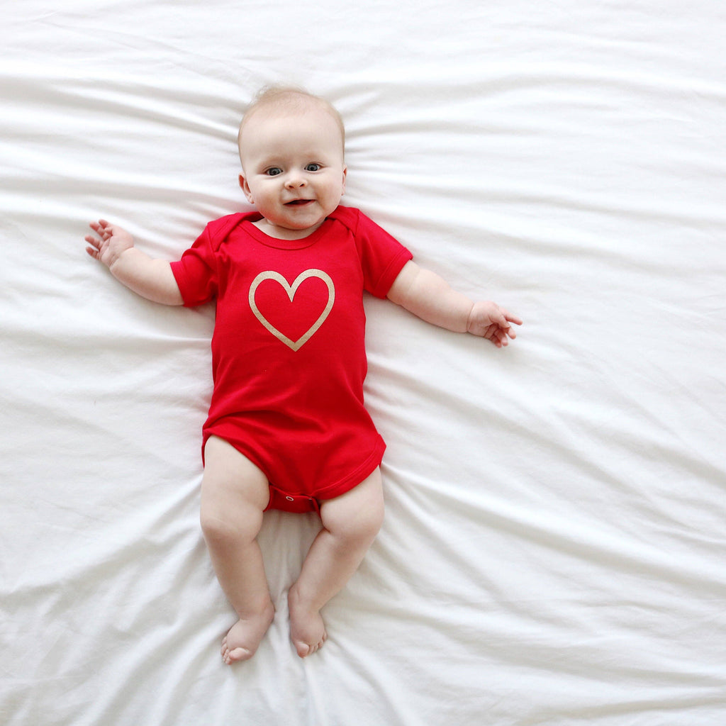 Baby wearing a red vest with a heart design on a white bed.