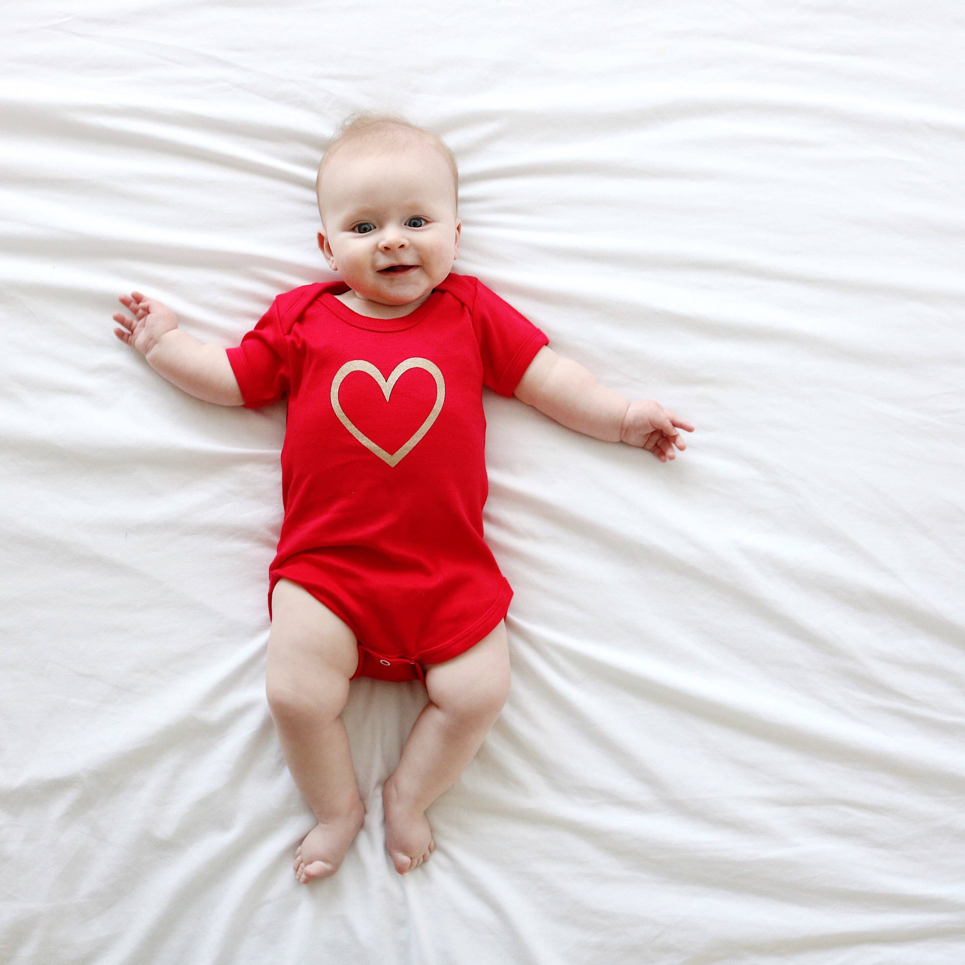 Baby wearing a red vest with a heart design on a white bed.