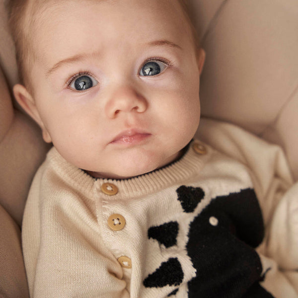 Baby wearing a beige sweater with black patterns, sitting on a soft surface.