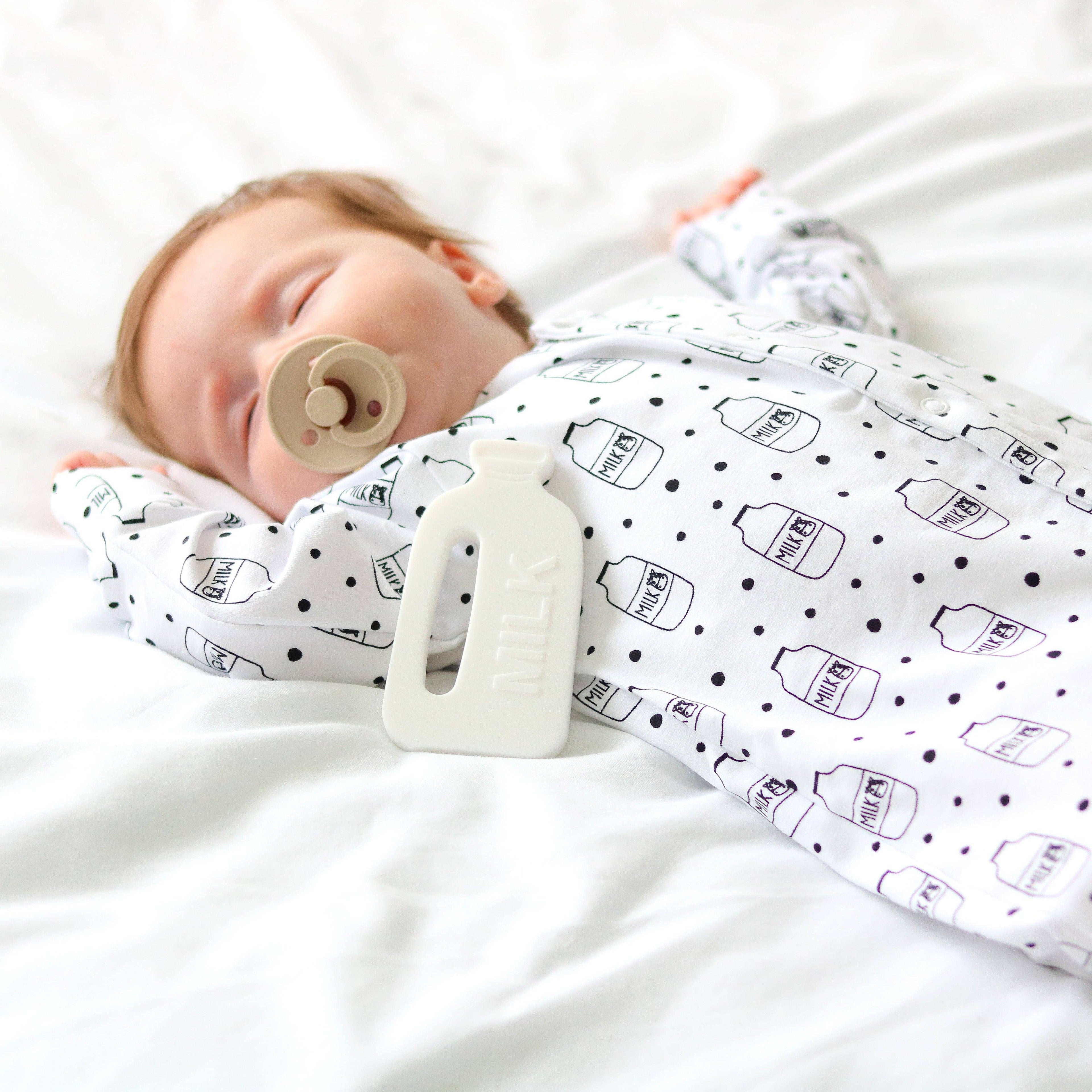 Baby wrapped in a sleepsuit with bottle patterns, lying on a white surface.