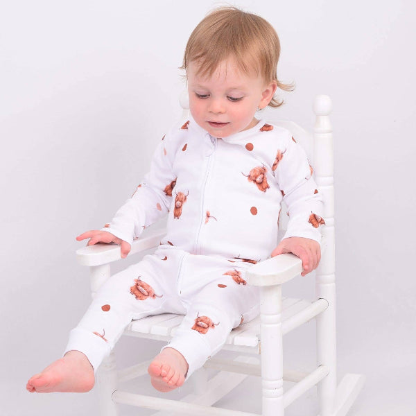 Child wearing a white outfit with highland cow patterns sitting on a white chair against a plain background