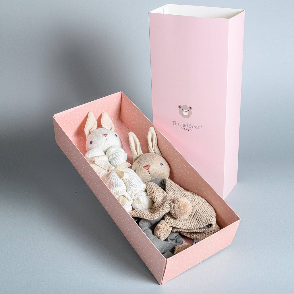 Two knitted rabbit toys in a pink box with a pink lid on a gray background