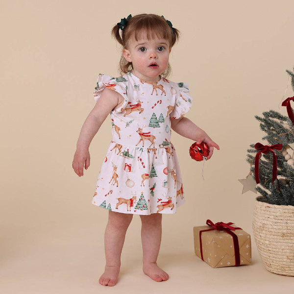 Child wearing a Christmas-themed dress with reindeer and trees, standing next to a small Christmas tree and gift box on a beige background.
