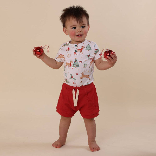 Child wearing a festive outfit with a white shirt and red shorts, holding Christmas ornaments on a beige background.