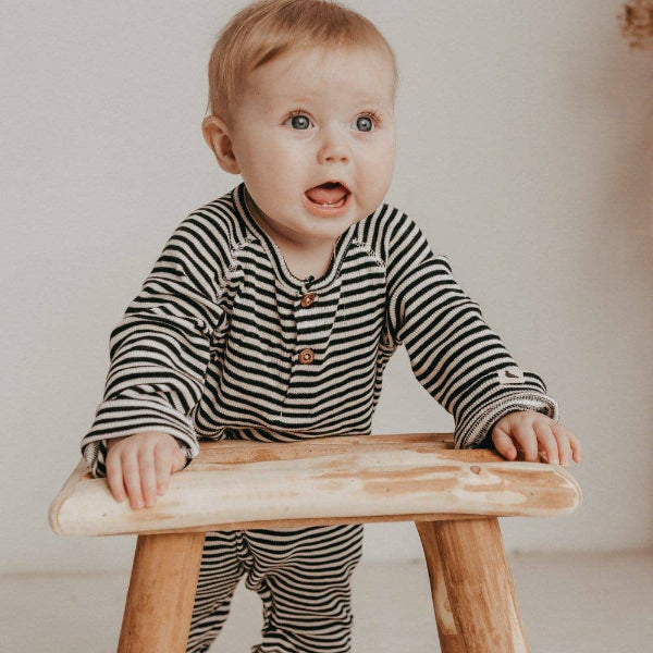 Baby in a striped rib playsuit sitting on a wooden stool against a plain background