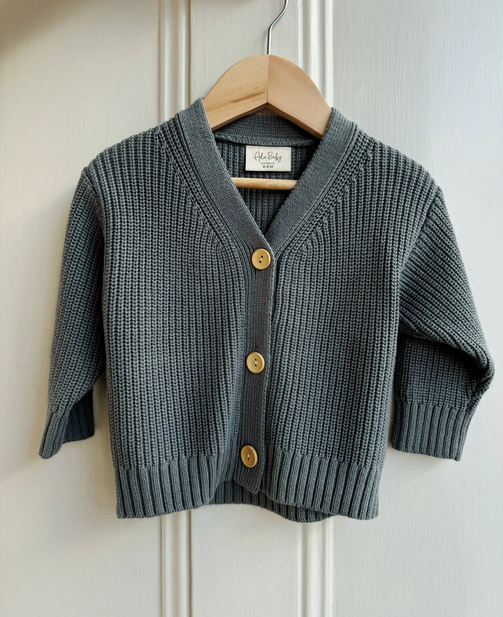 Deep dark green knitted cardigan with gold buttons on a wooden hanger against a white wall.