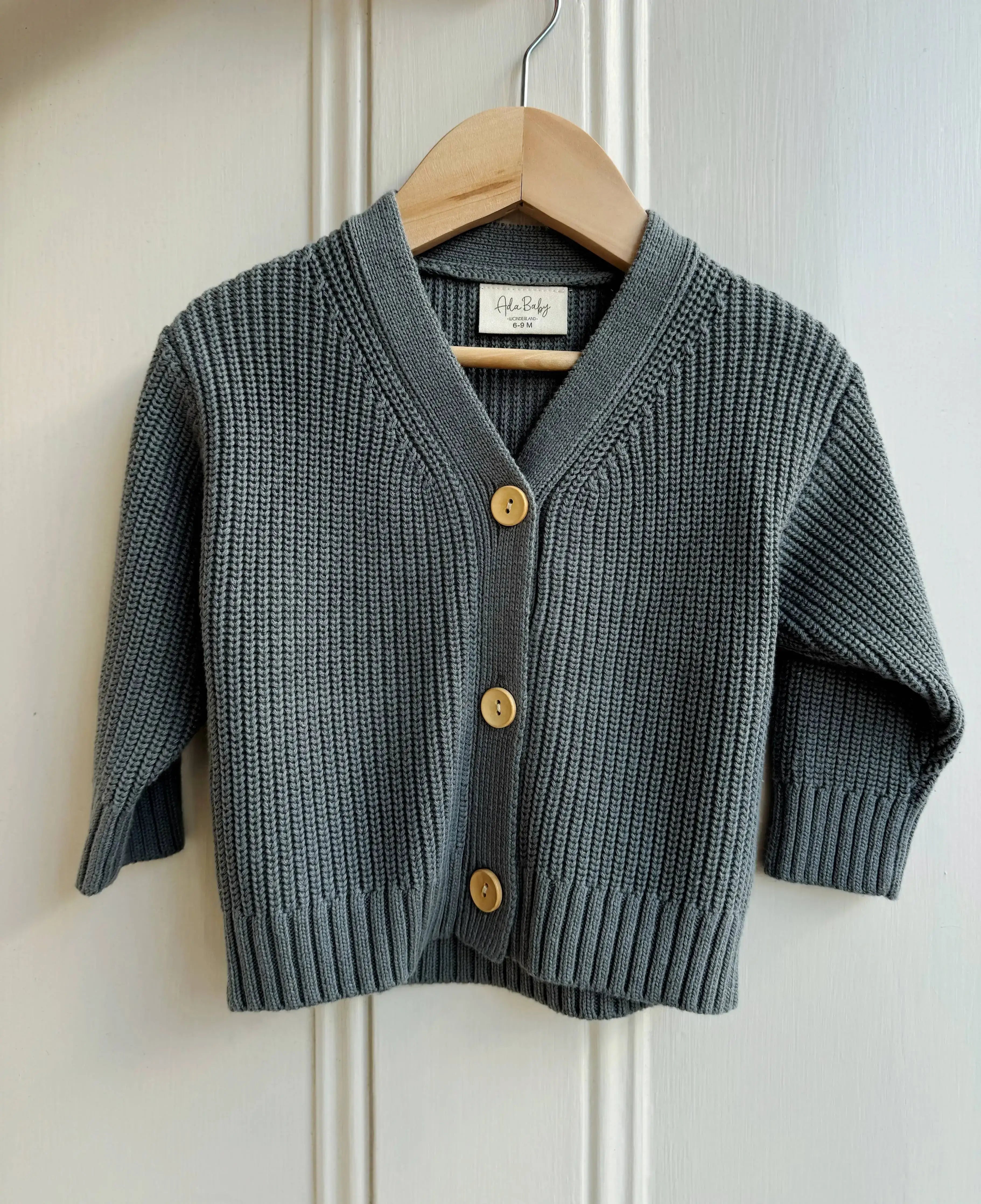 Deep dark green knitted cardigan with gold buttons on a wooden hanger against a white wall.