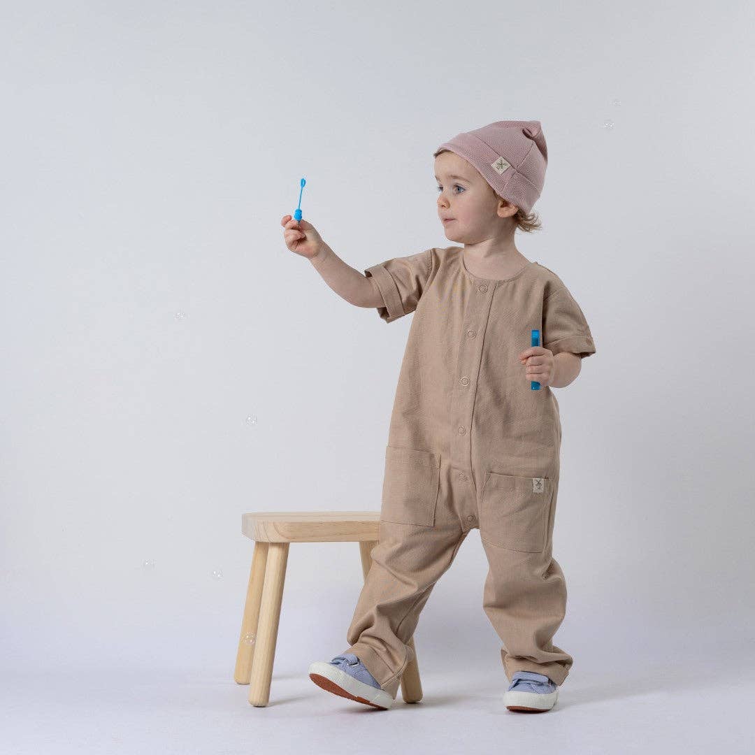 Child wearing a beige onesie and pink hat, standing next to a small wooden stool on a white background.