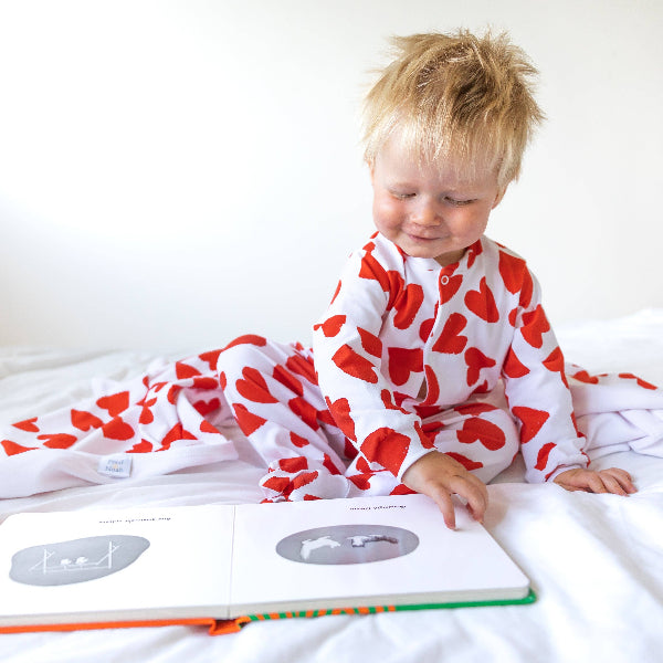 Child wearing a red and white patterned onesie, sitting on a bed with a book open in front of them.