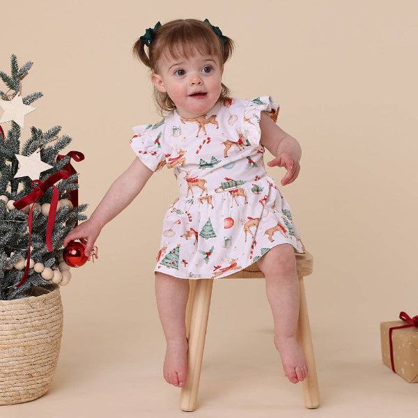 Child in a Christmas-themed dress standing next to a decorated tree on a beige background