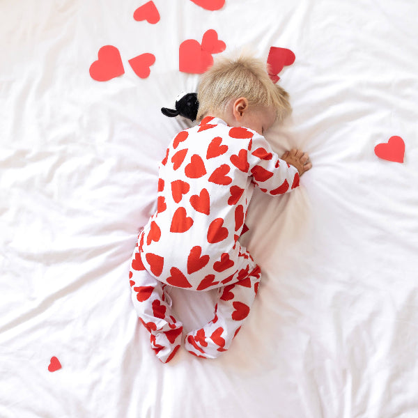 Baby wearing a red heart-patterned onesie lying on a white surface with scattered red hearts.