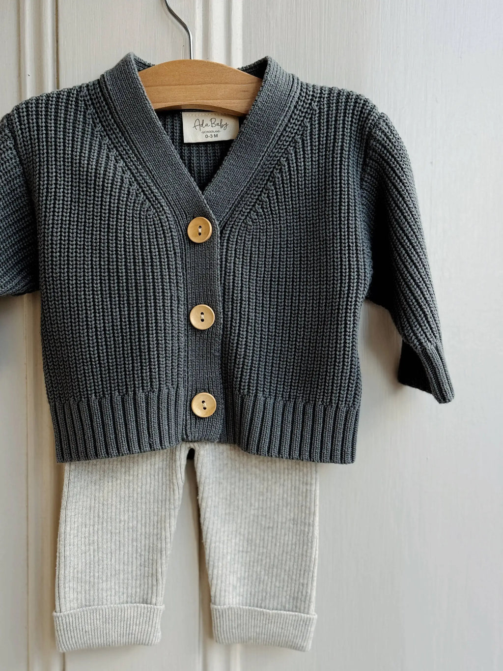 Children's deep dark green cardigan with wooden buttons and white pants on a hanger against a white wall.