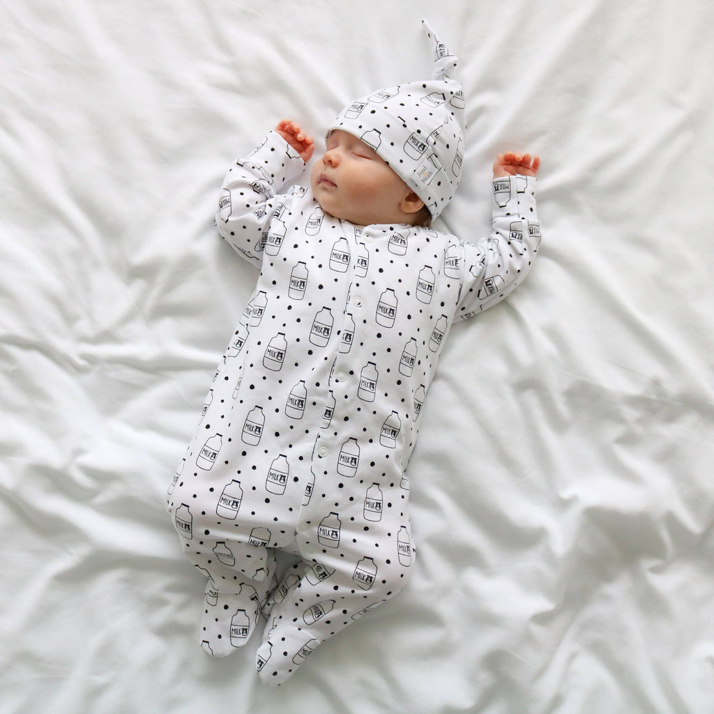 Newborn baby wearing a white sleepsuit with black patterns and a matching hat, lying on a white bed.