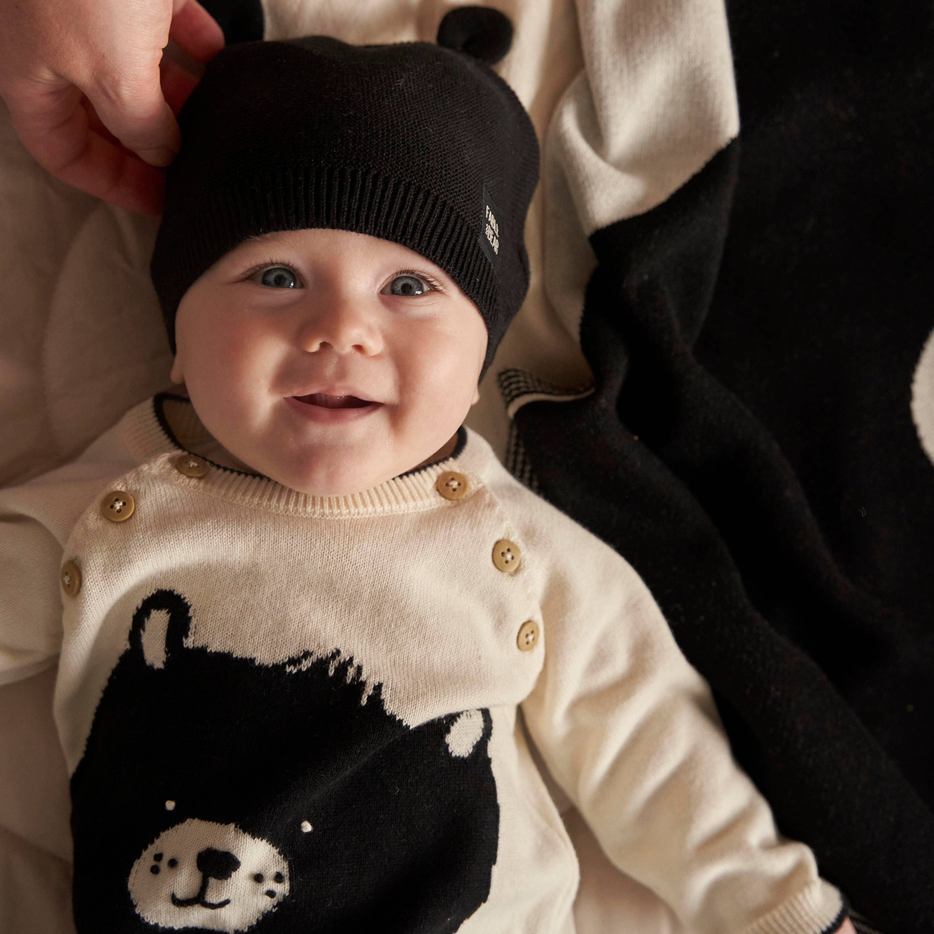 Baby wearing a black knit hat and a cream jumper/sweater with a bear design.
