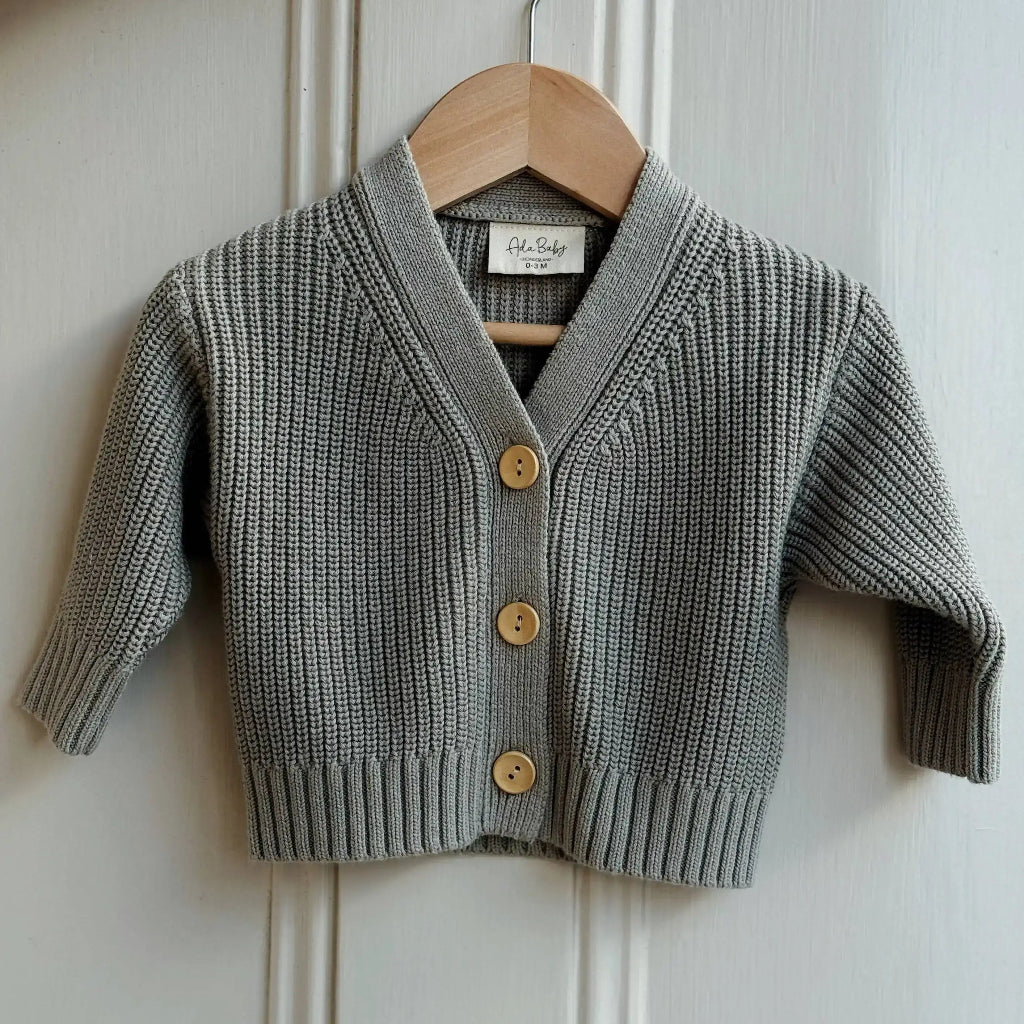 Light green knitted cardigan with yellow buttons on a wooden hanger against a white paneled wall.