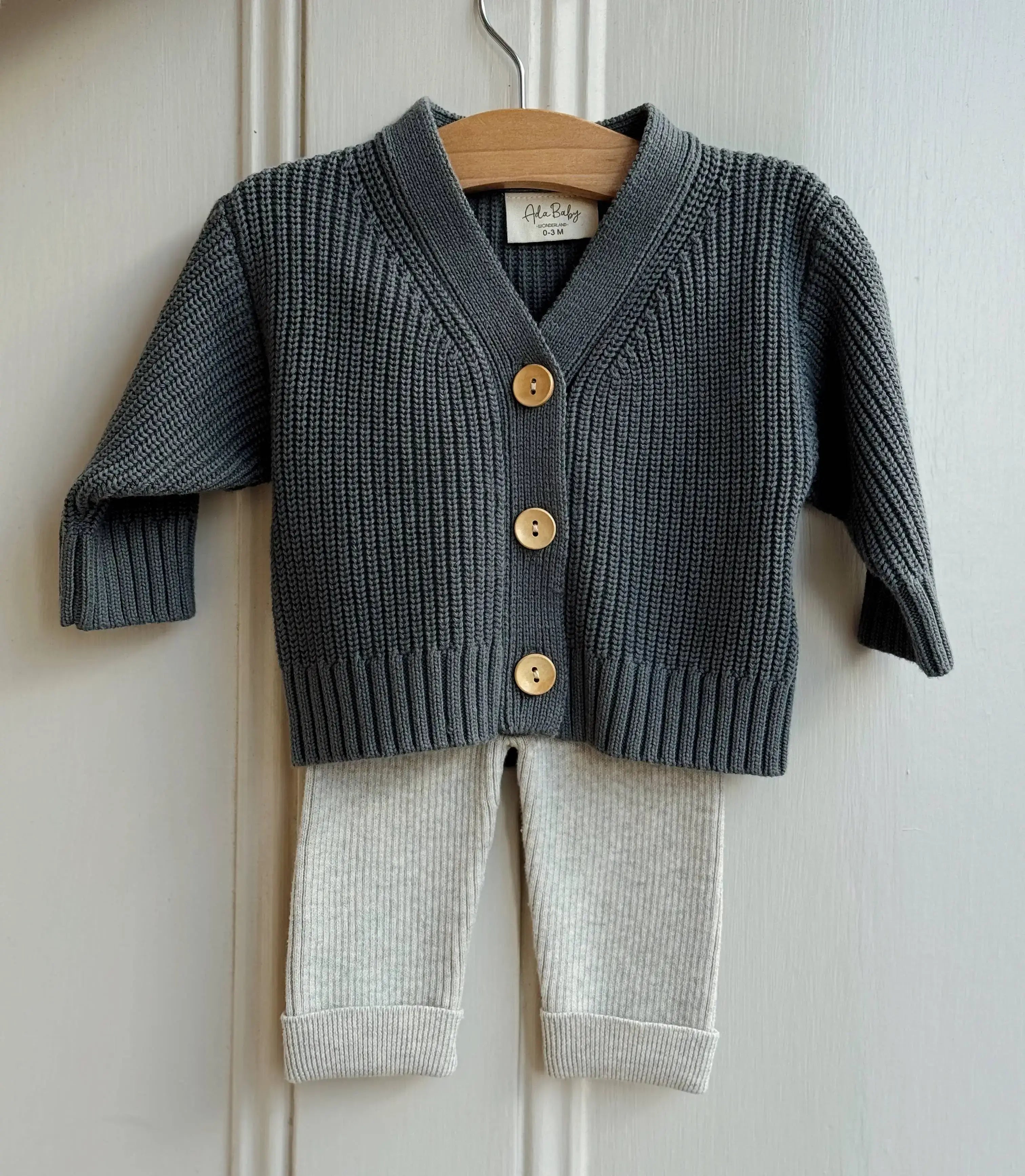Children's deep dark green knit cardigan with gold buttons and matching pants on a hanger against a white wall.
